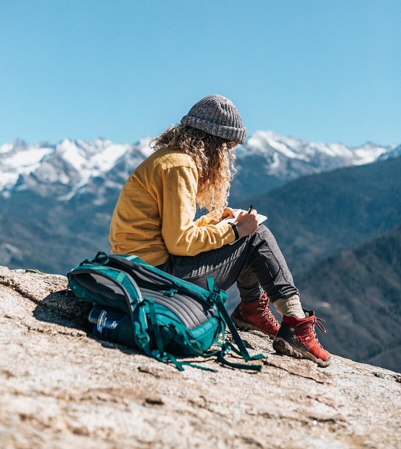 Woman dressed in mountain clothes, sat on mountain top taking notes with backpack beside her