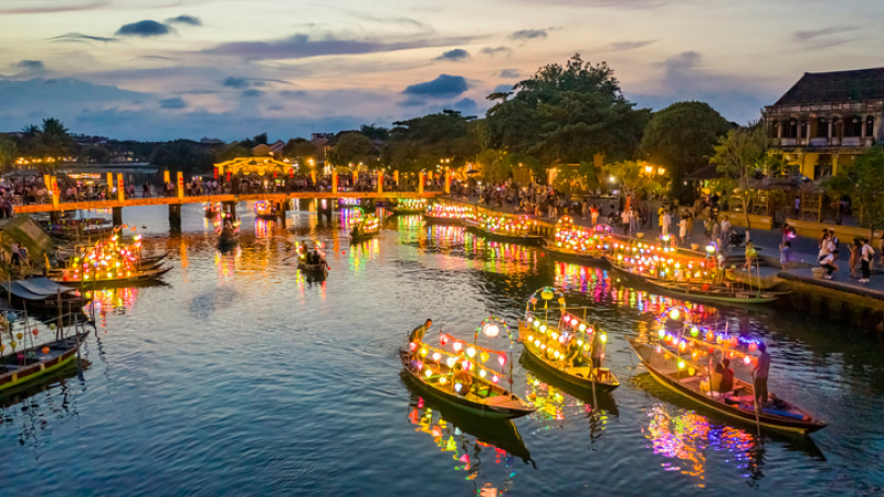 An aerial view of boats on a river at dusk. Multicoloured lights illuminate the boats and the water around them. A bridge across the water is lined with tourists and other onlookers.