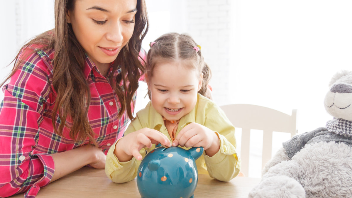 Mother overlooking her daughter placing a coin into a piggybank; both sat at a table
