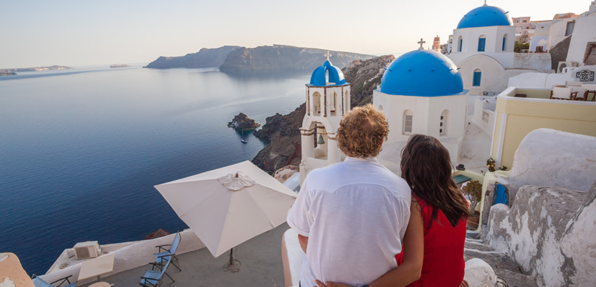 Couple sat embraced looking over the white and blue buildings of Greece along a rocky coastline 