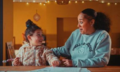 A child holding a pencil sits at a table with an adult wearing a 'SMELL-A-LICIOUS' sweatshirt