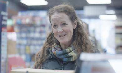 A person standing at the counter in a brightly lit Post Office branch, with shelves of products in the background