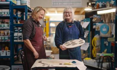 Two people in a workshop wearing aprons, one holding a tray of small clay objects, surrounded by shelves filled with tools and supplies