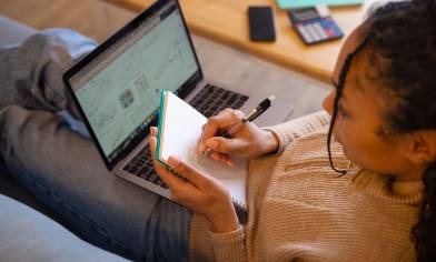 Person sitting on a sofa writing in a notebook while using a laptop, with charts visible on the screen; a smartphone, calculator, and folder are on the table nearby.