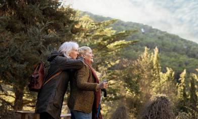 Two people stand together in a forested area, with one person hugging the other from behind while the other holds a walking stick
