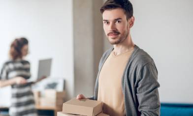 Man holding cardboard parcels and smiling at the camera, with a woman in the background looking at a laptop in a home setting.