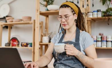 A person sitting at a table with a laptop, holding a mug, surrounded by various pottery items and plants