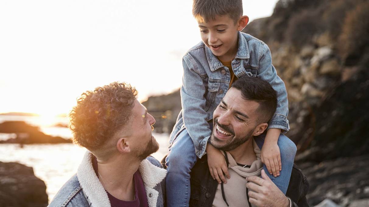A happy family moment showing two men smiling with a young boy sitting on one man’s shoulders, outdoors near a rocky coastline