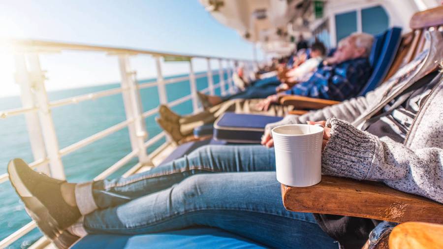 A peaceful moment at sea. A group of people lounge on deck chairs aboard a cruise ship, soaking up the sun and gazing out at the open ocean, with one person in the foreground holding a white cup.
