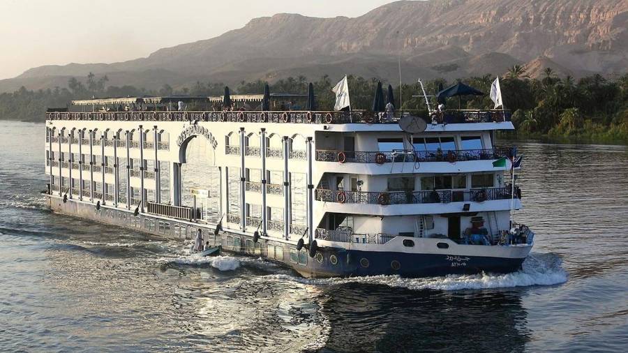 A river cruise ship sails past greenery and distant mountains