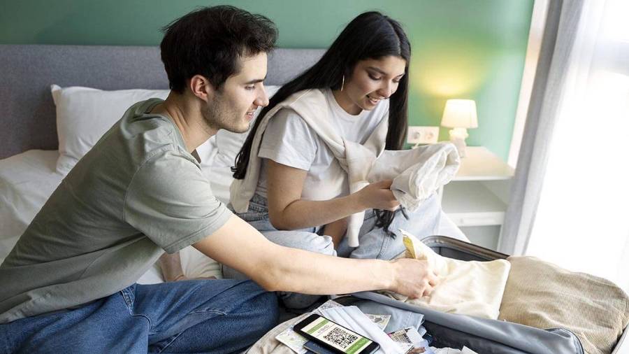Two people sit on a bed with an open suitcase containing clothes, a passport, travel documents and a smartphone displaying a QR code, with a bedside table and lamp in the background