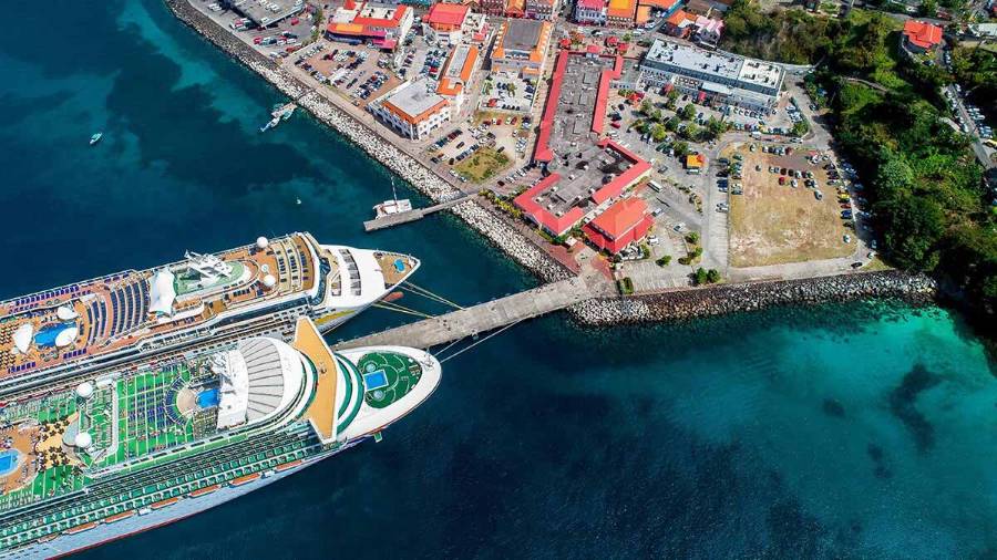 Aerial view of a coastal area and developed port area with two large cruise ships docked at a pier, surrounded by clear blue water