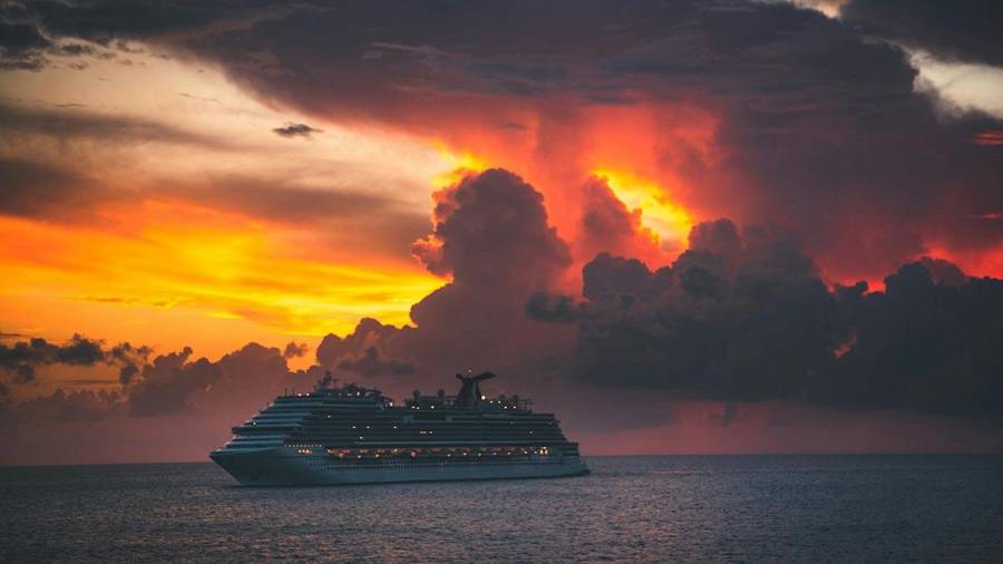 A cruise ship sails on calm water beneath a vivid sunset with glowing clouds