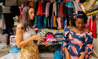 Two individuals standing in a clothing market with colourful garments.