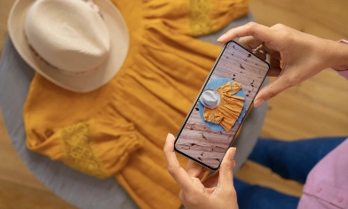 A person holding a smartphone is taking a photo of a yellow dress and a beige hat placed on a round table.