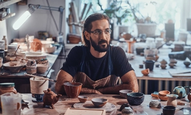 A man sitting at a workbench in a pottery studio with various pottery tools, brushes, and several pieces of pottery in different stages of completion.