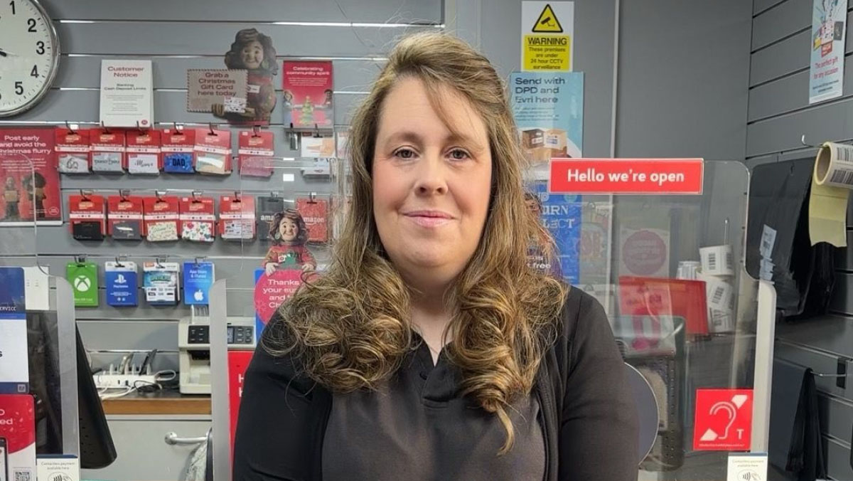 Helen, a postmaster, with long wavy hair, wearing a black top, standing in front of a display of gift cards and signage in her Post Office branch.