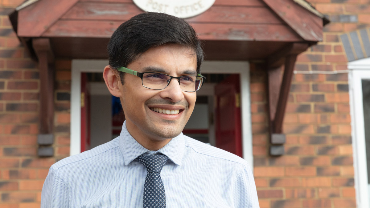 Dhaval, a postmaster, wearing glasses, a light blue shirt and a patterned tie. He is smiling warmly in front of his Post Office branch.