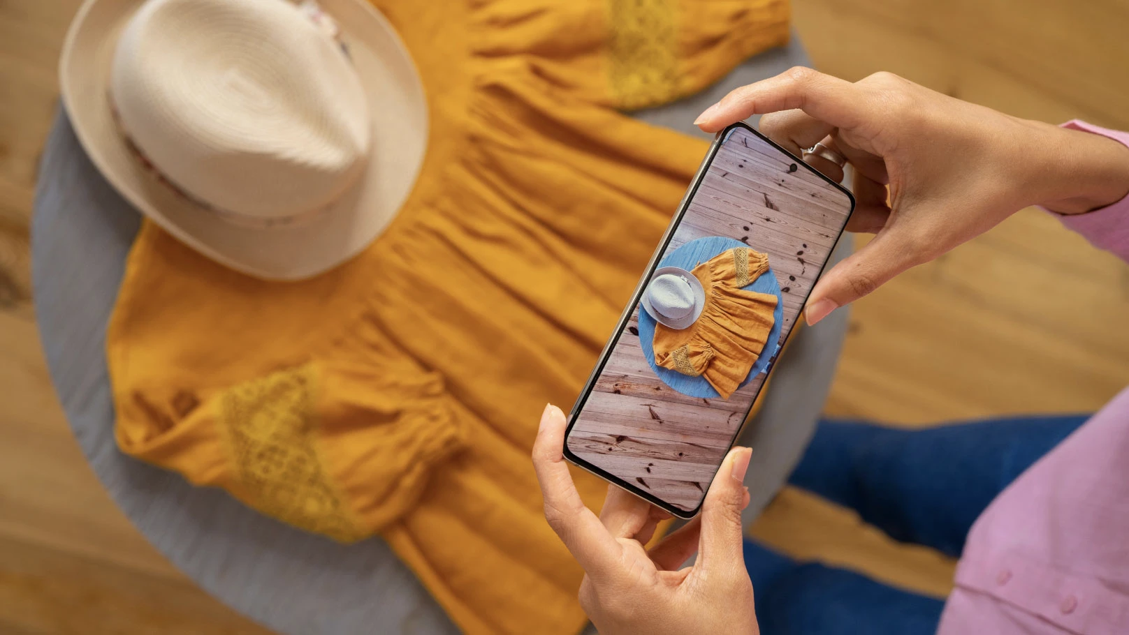 A person holding a smartphone is taking a photo of a yellow dress and a beige hat placed on a round table.