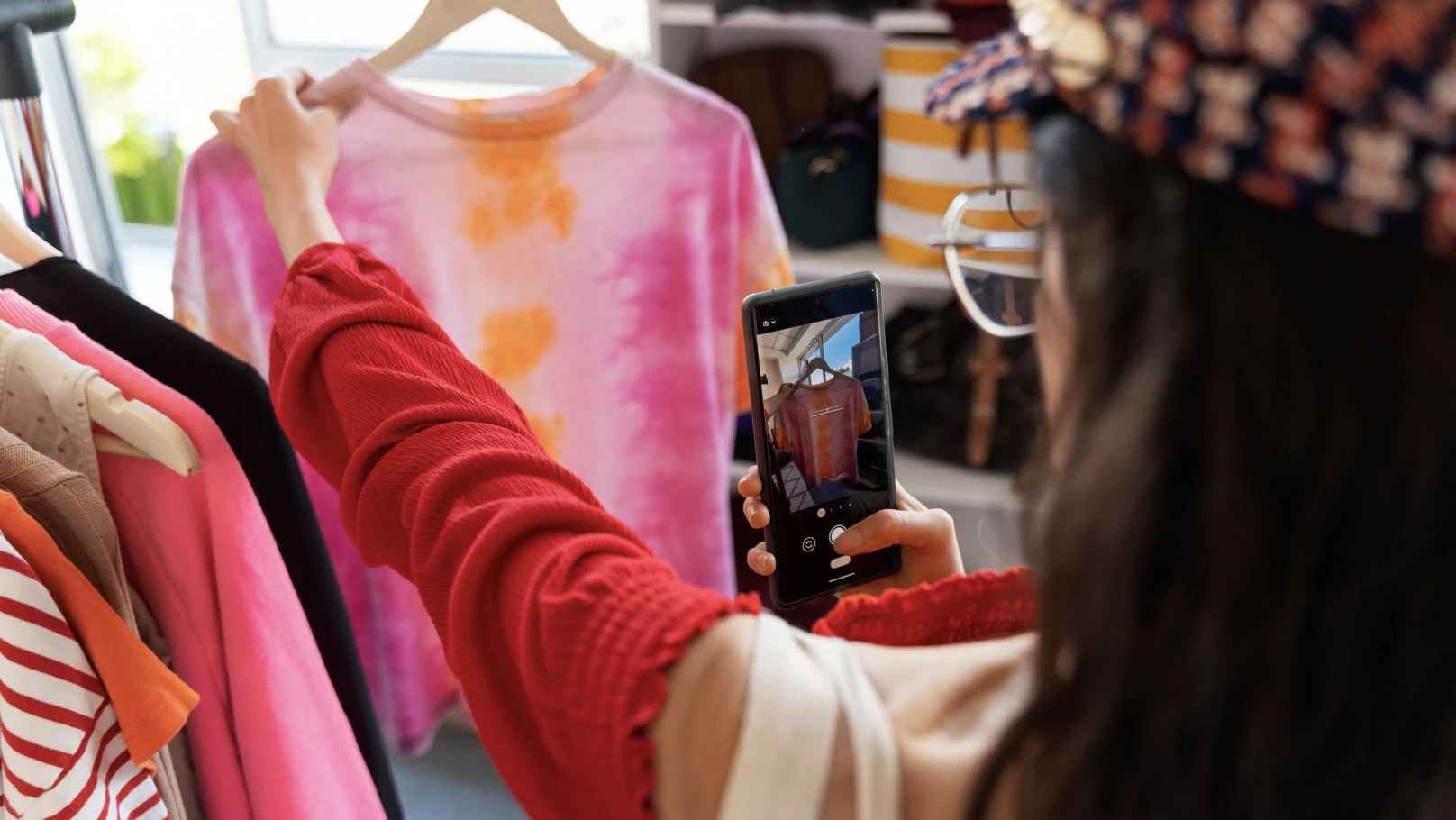 A person holding a tie-dye shirt with pink and orange patterns on a hanger and taking a photo of it with a smartphone.