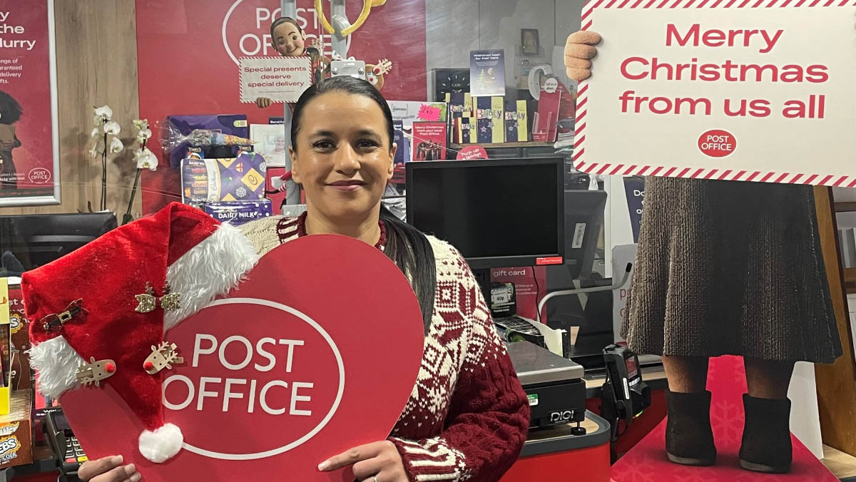 Gurpreet, a postmaster, in a festive jumper, holding a heart-shaped Post Office board with a Santa hat in a Christmas-themed branch.