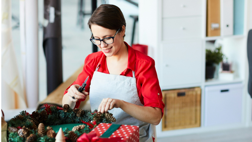 An individual crafting a holiday wreath at a table covered with festive decorations