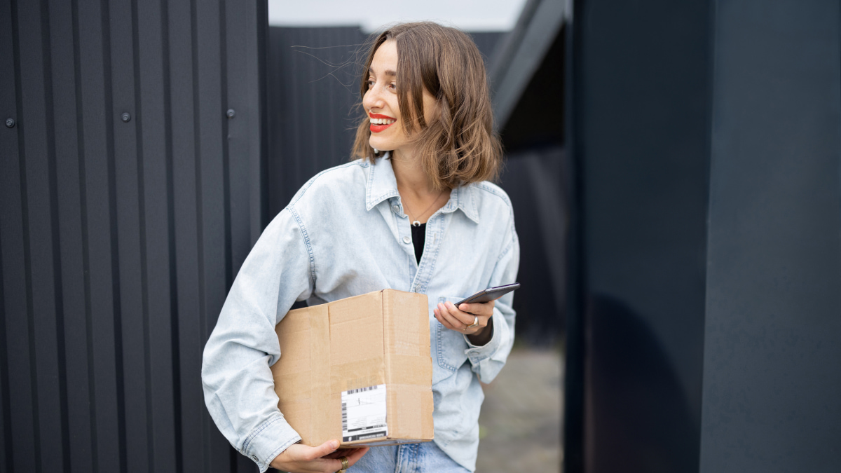 Person in a denim shirt holding a cardboard package and a smartphone while standing in front of a black corrugated fence next to an open gate.