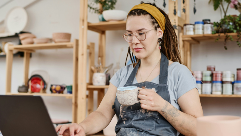 A person sitting at a table with a laptop, holding a mug, surrounded by various pottery items and plants 